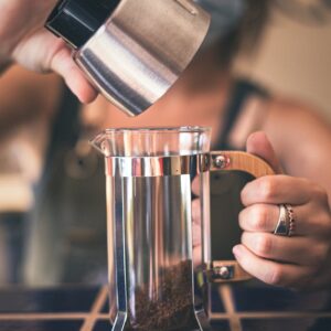 Barista adding coffee grounds to a French press in a café setting.